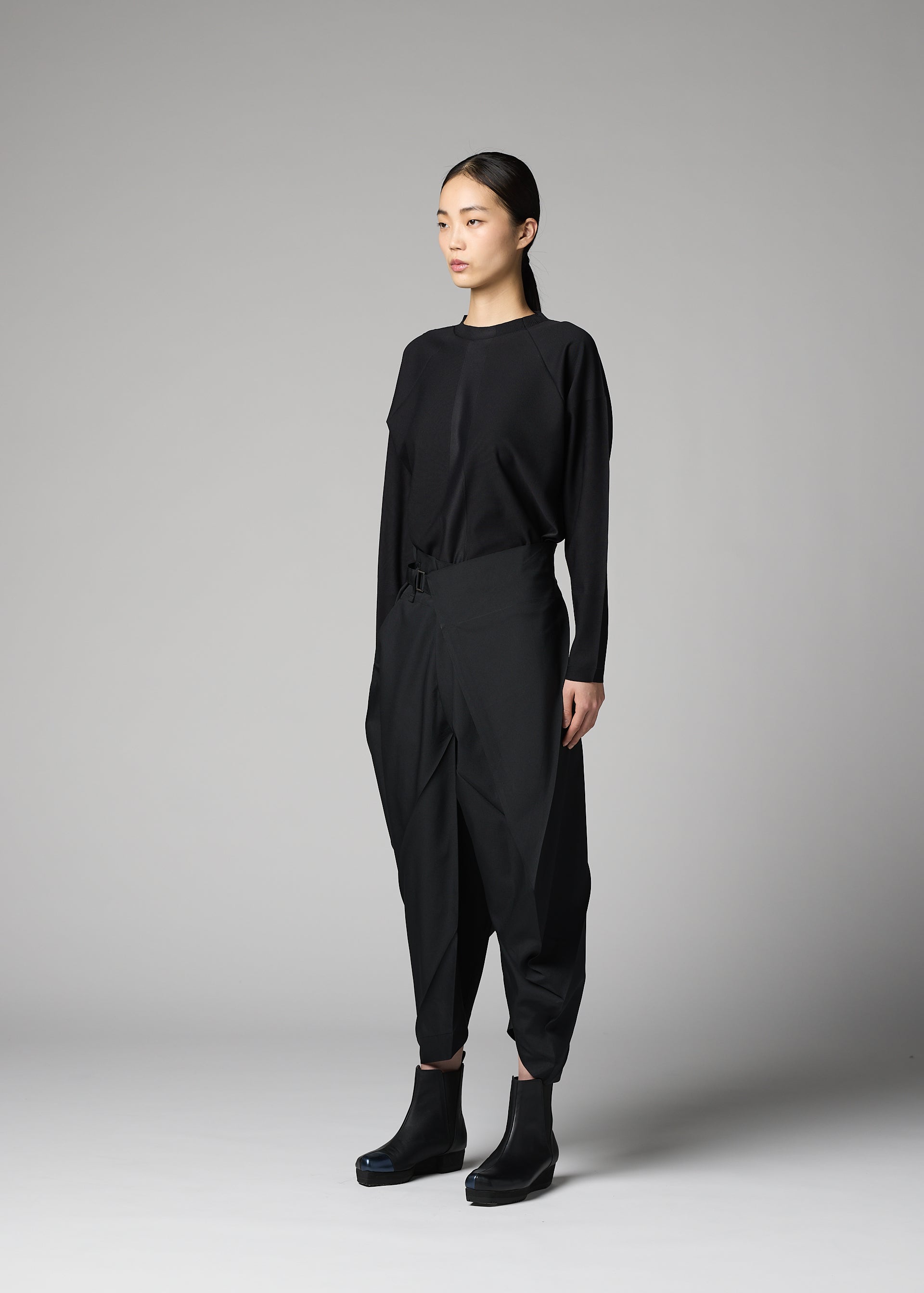 Zoomed view A person stands against a gray background, wearing the F-BT pants by 132 5. ISSEY MIYAKE—black folding technique trousers paired with a black long-sleeve top and platform ankle boots. Their straight dark hair is tied back as they look slightly to the side.