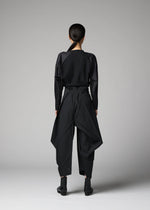 A person with long dark hair in a low ponytail stands facing away, wearing the F-BT wide-legged pants with draped panels by 132 5. ISSEY MIYAKE, a black long-sleeve top, and black ankle boots against a plain gray background.