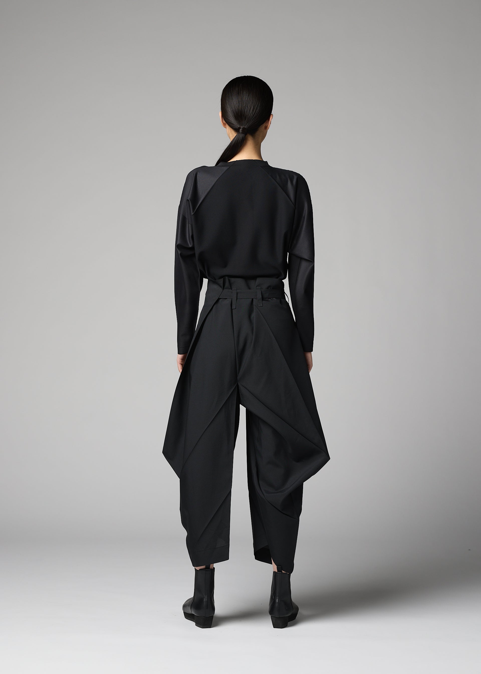 Zoomed view A person with long dark hair in a low ponytail stands facing away, wearing the F-BT wide-legged pants with draped panels by 132 5. ISSEY MIYAKE, a black long-sleeve top, and black ankle boots against a plain gray background.