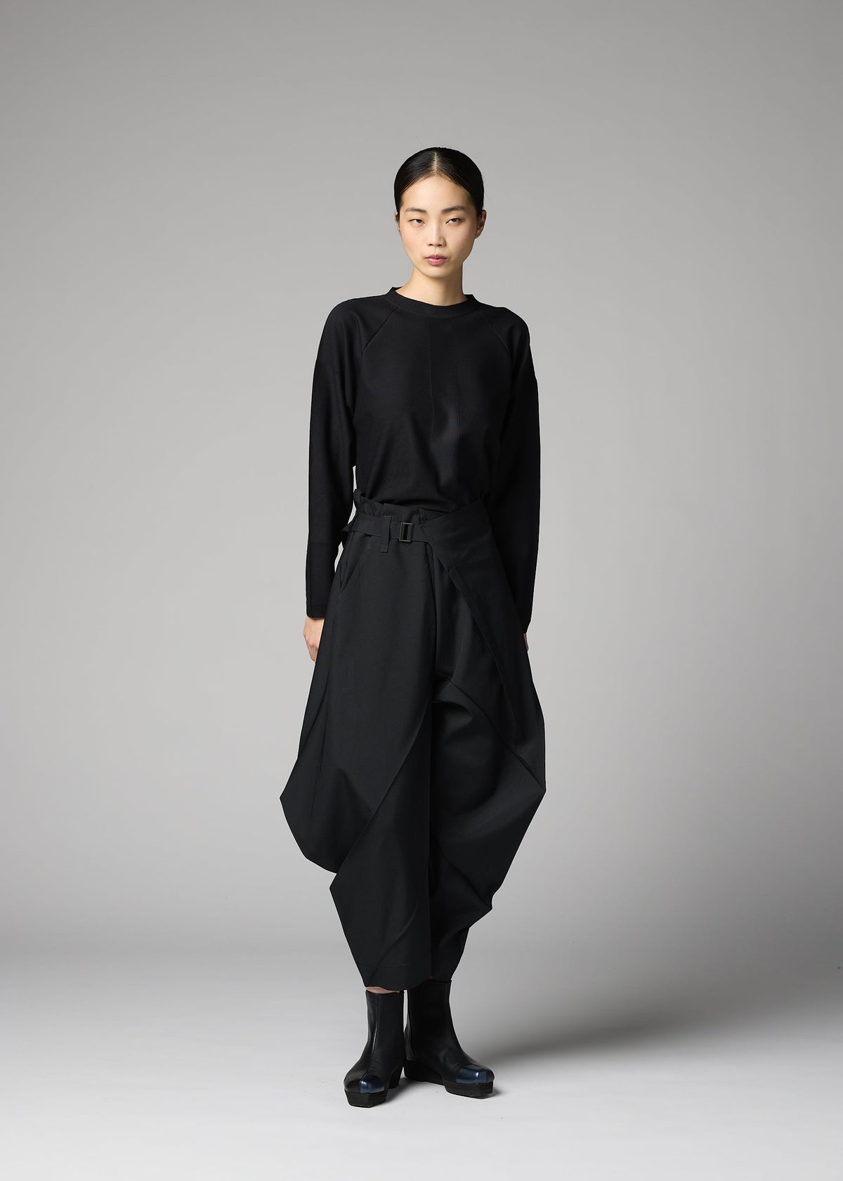 A person stands against a plain gray background, wearing 132 5. ISSEY MIYAKE's F-BT black long-sleeve top and solid color folding technique pants made from recycled polyester, paired with black ankle boots. Their hair is pulled back; their expression is neutral. view 9 of 9