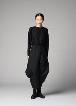 A person stands against a plain gray background, wearing 132 5. ISSEY MIYAKE's F-BT black long-sleeve top and solid color folding technique pants made from recycled polyester, paired with black ankle boots. Their hair is pulled back; their expression is neutral.