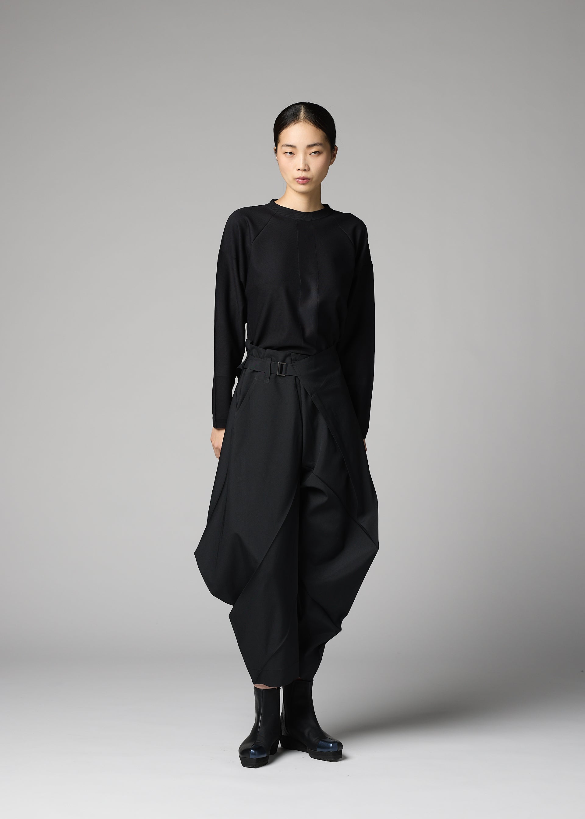 Zoomed view A person stands against a plain gray background, wearing 132 5. ISSEY MIYAKE's F-BT black long-sleeve top and solid color folding technique pants made from recycled polyester, paired with black ankle boots. Their hair is pulled back; their expression is neutral.