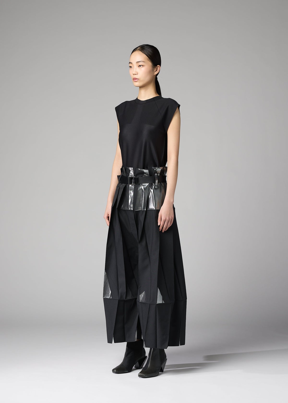 A woman poses in a studio against a plain gray background, wearing No.3-1 by 132 5. ISSEY MIYAKE—an innovative black outfit featuring geometric panels, metallic accents, foil-detailed pants, and black ankle boots. view 9 of 10