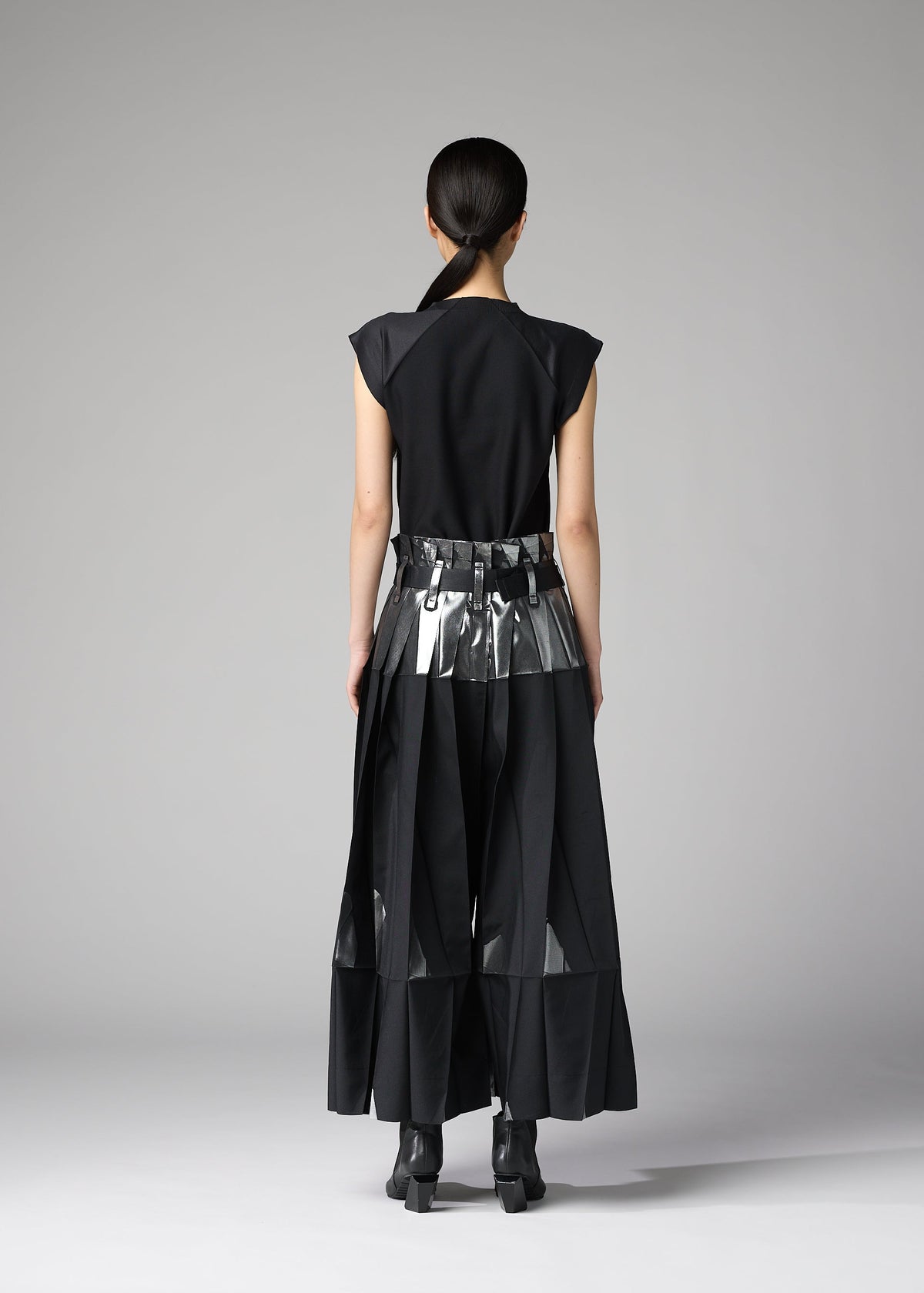 A person faces away in a minimal studio, wearing the No.3-1 set by 132 5. ISSEY MIYAKE: a sleek black outfit with foil-detailed pants and a metallic belt over a long skirt; their hair is tied back. view 10 of 10