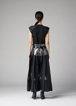 A person faces away in a minimal studio, wearing the No.3-1 set by 132 5. ISSEY MIYAKE: a sleek black outfit with foil-detailed pants and a metallic belt over a long skirt; their hair is tied back.