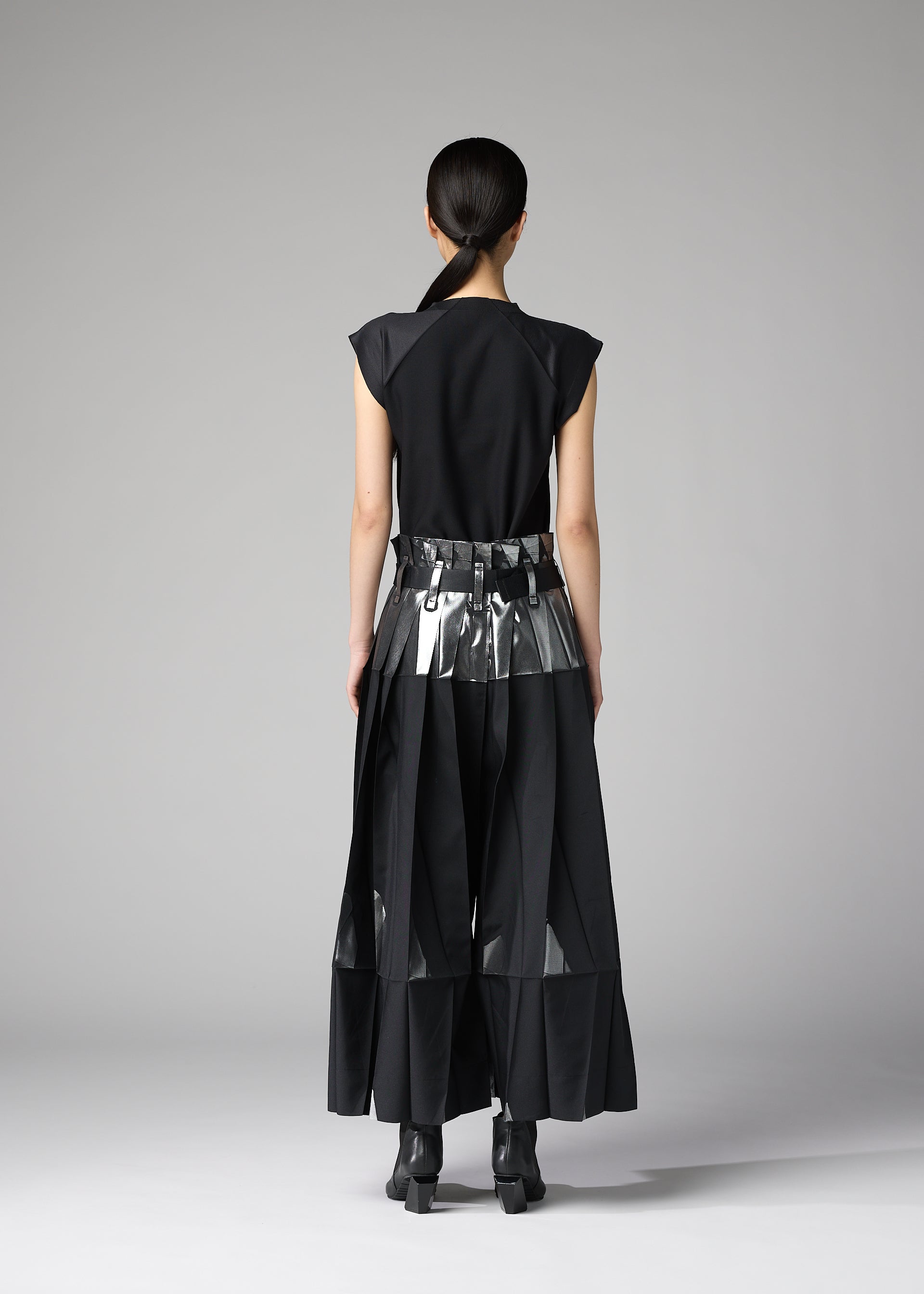 Zoomed view A person faces away in a minimal studio, wearing the No.3-1 set by 132 5. ISSEY MIYAKE: a sleek black outfit with foil-detailed pants and a metallic belt over a long skirt; their hair is tied back.