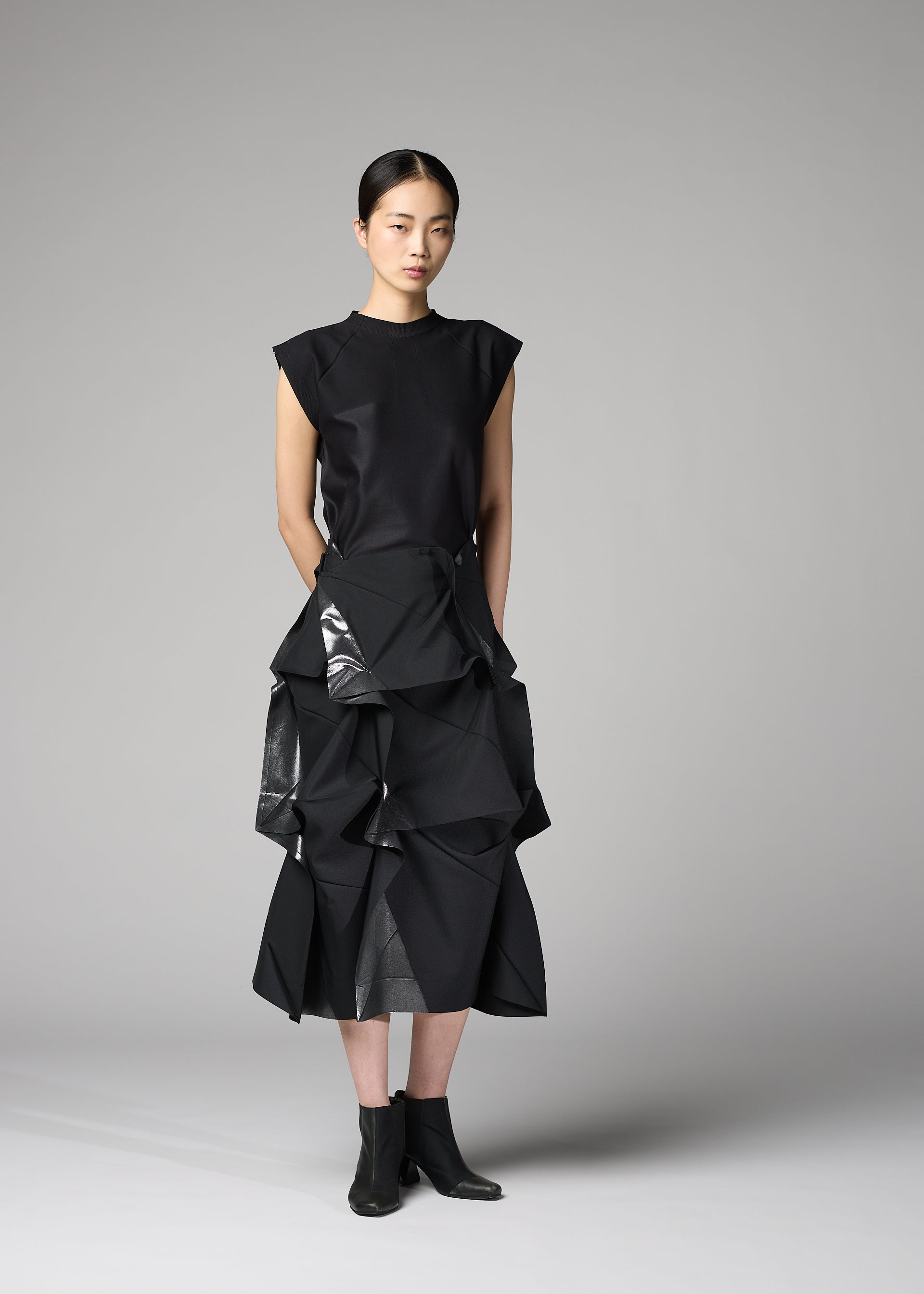 Zoomed view A woman stands in a studio wearing the No.1-2 skirt from 132 5. ISSEY MIYAKE—a sleeveless black top paired with a layered, ruffled black foil skirt featuring shiny accents and a flared silhouette, completed with black ankle boots. Her straight dark hair is pulled back, and she maintains a neutral expression.