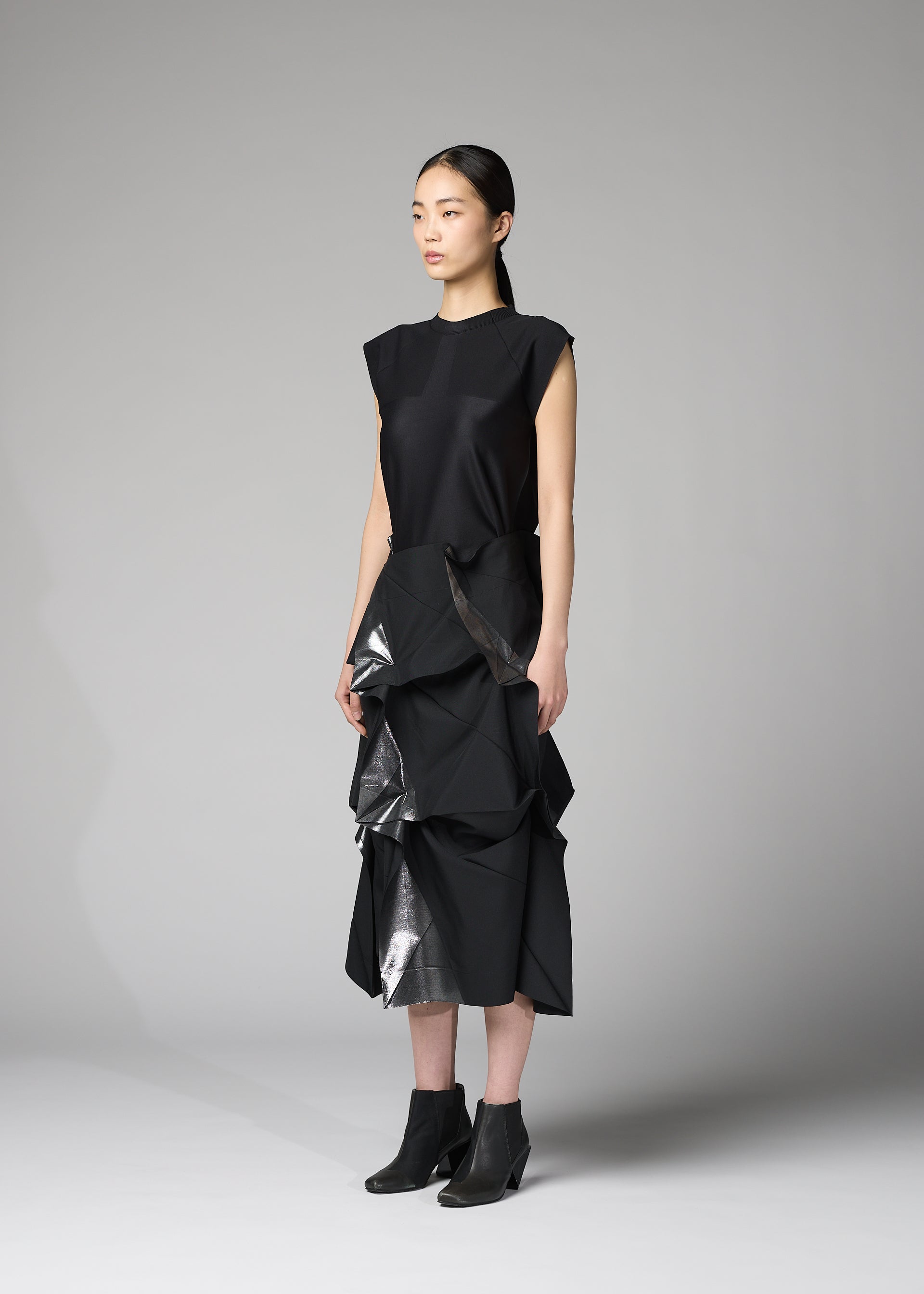 Zoomed view A person stands in a minimalist studio wearing the No.1-2 skirt by 132 5. ISSEY MIYAKE—a recycled polyester piece featuring shiny, sculptural ruffles in a flared silhouette—styled with a sleeveless black top and black ankle boots.