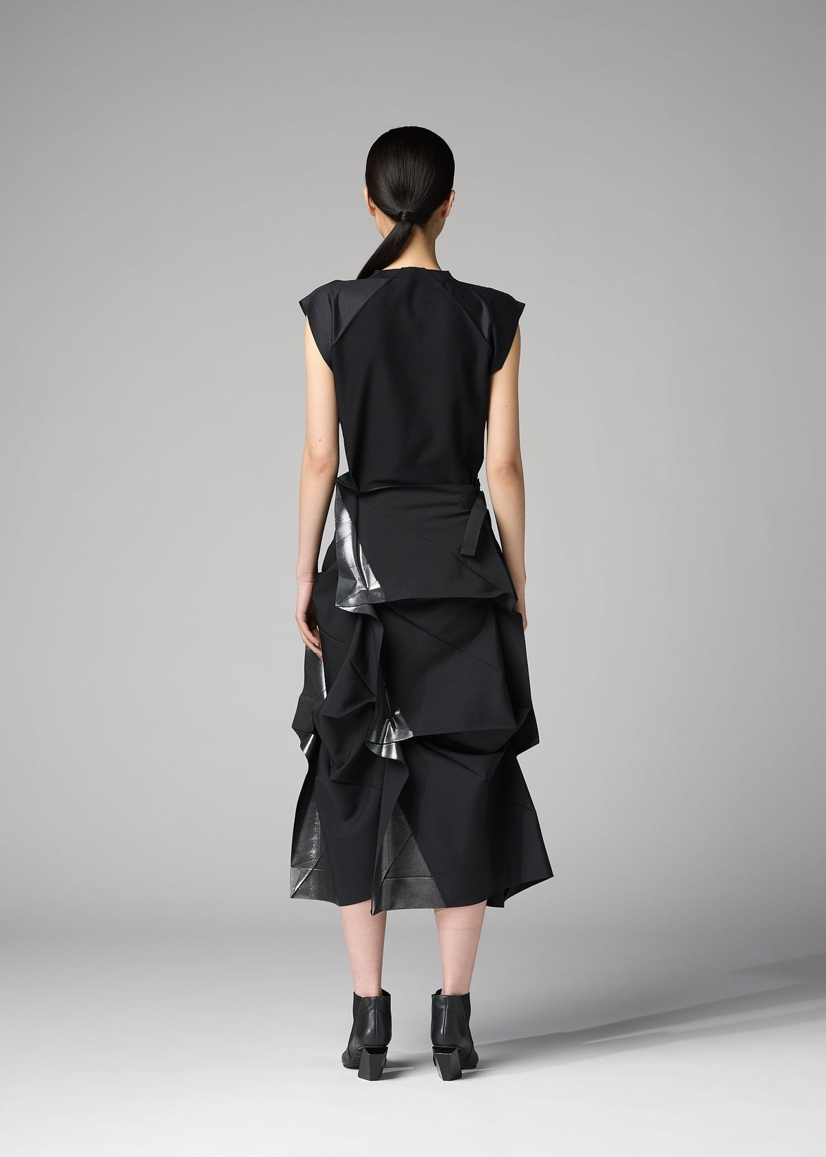 A woman stands facing away, wearing the 132 5. ISSEY MIYAKE No.1-2 black dress with a flared silhouette and layered, angular details, paired with black ankle boots. Her hair is tied back in a low ponytail against a plain gray background. view 8 of 10