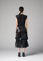 A woman stands facing away, wearing the 132 5. ISSEY MIYAKE No.1-2 black dress with a flared silhouette and layered, angular details, paired with black ankle boots. Her hair is tied back in a low ponytail against a plain gray background.