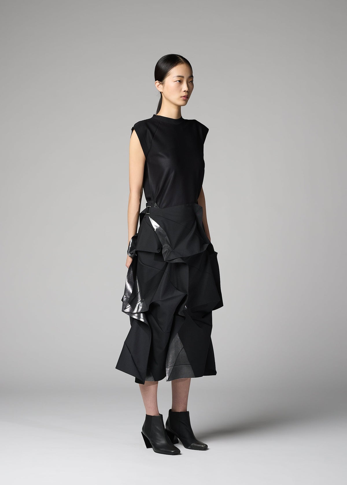 A person stands in a studio wearing the No.1-2 by 132 5. ISSEY MIYAKE: a sleeveless black top and a layered, sculptural black skirt with shiny accents reminiscent of foil, paired with pointed black ankle boots. The plain gray background highlights the look. view 9 of 10