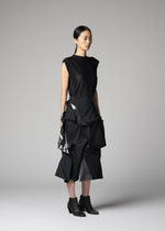 A person stands in a studio wearing the No.1-2 by 132 5. ISSEY MIYAKE: a sleeveless black top and a layered, sculptural black skirt with shiny accents reminiscent of foil, paired with pointed black ankle boots. The plain gray background highlights the look.