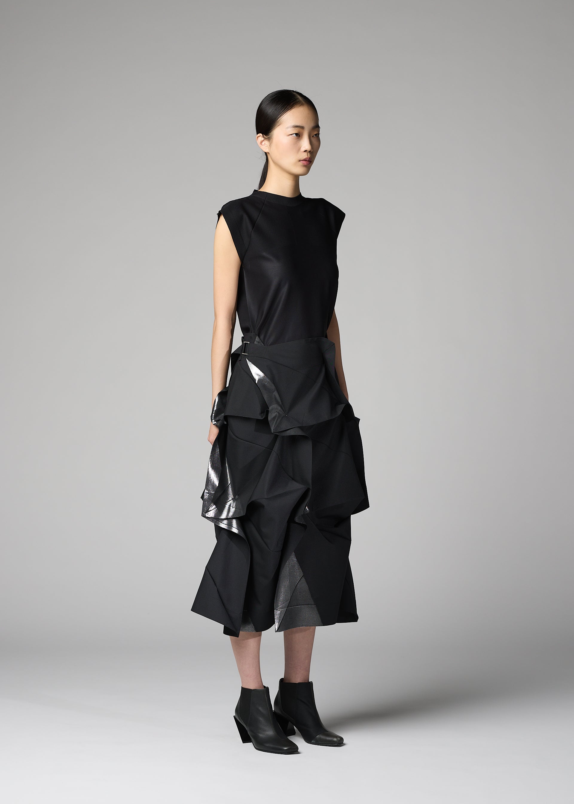 Zoomed view A person stands in a studio wearing the No.1-2 by 132 5. ISSEY MIYAKE: a sleeveless black top and a layered, sculptural black skirt with shiny accents reminiscent of foil, paired with pointed black ankle boots. The plain gray background highlights the look.