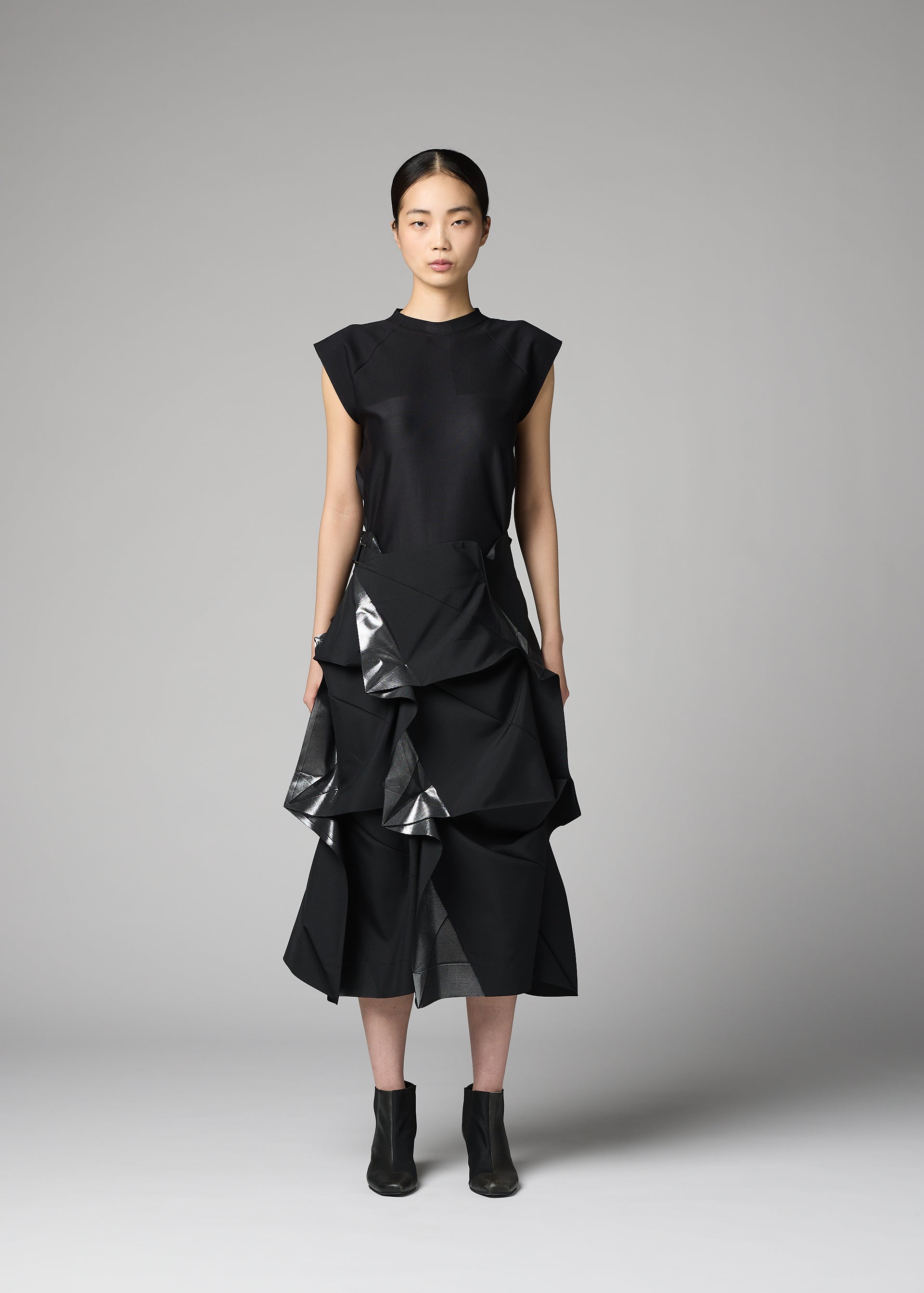 Zoomed view A woman stands against a gray backdrop wearing the 132 5. ISSEY MIYAKE No.1-2—a black sleeveless top paired with a recycled polyester skirt featuring geometric, structured ruffles that create a flared silhouette. She completes the look with black ankle boots, facing forward with a neutral expression.