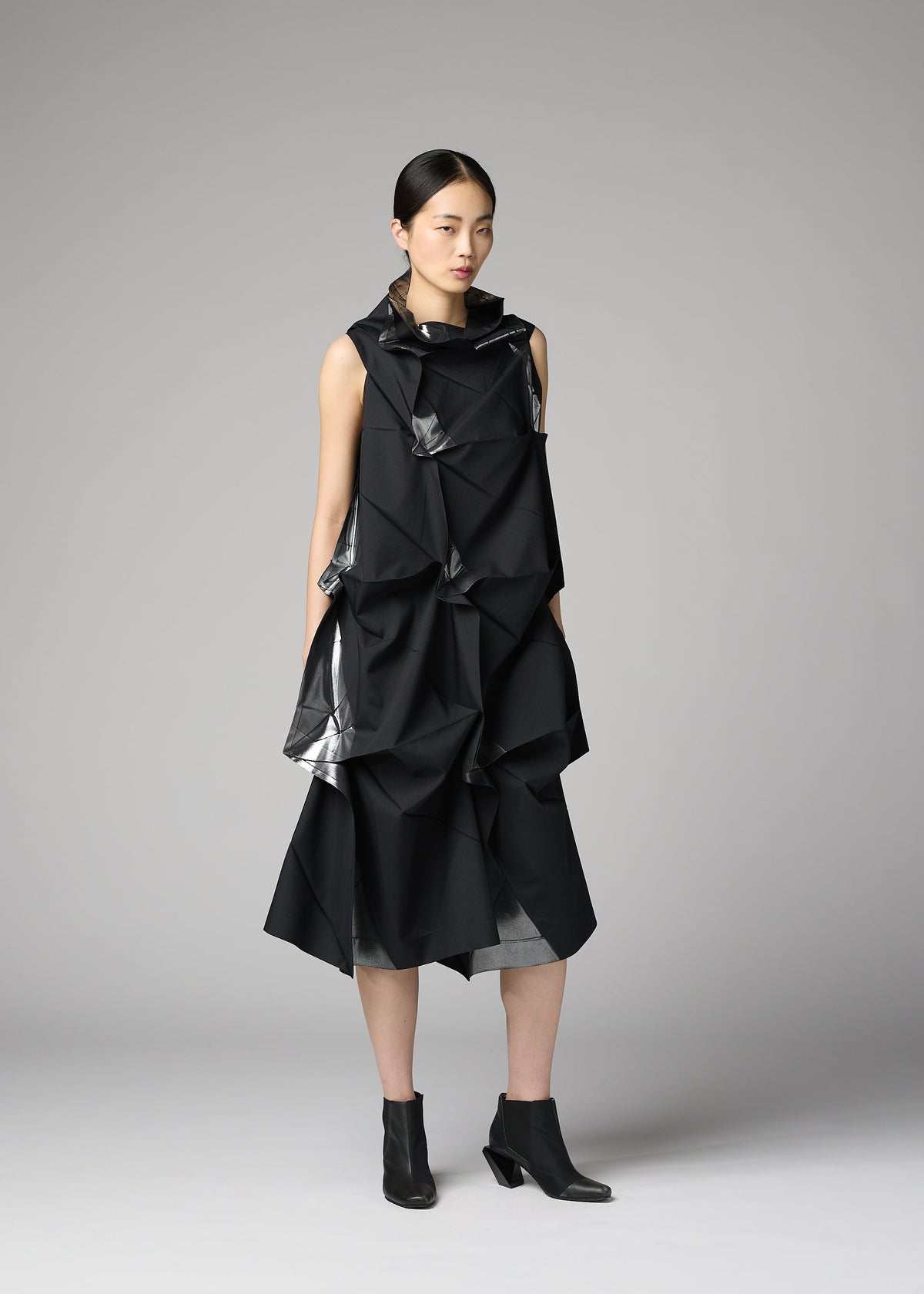 A woman poses in a studio against a neutral gray backdrop, wearing 132 5. ISSEY MIYAKE’s No.1-1—a sleeveless black recycled polyester dress with angular folds and shiny accents—paired with black ankle boots. view 5 of 9