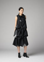A woman poses in a studio against a neutral gray backdrop, wearing 132 5. ISSEY MIYAKE’s No.1-1—a sleeveless black recycled polyester dress with angular folds and shiny accents—paired with black ankle boots.