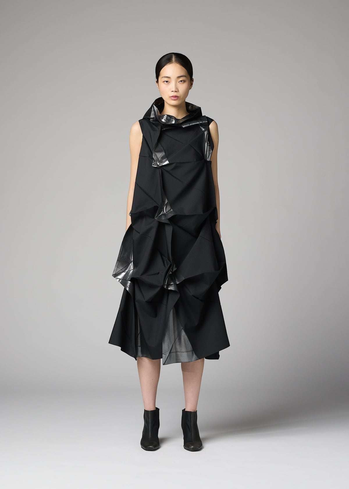 A woman with straight dark hair and a neutral expression stands against a plain gray background, wearing 132 5. ISSEY MIYAKE’s No.1-1: a sleeveless black recycled polyester dress with origami-like folds, shiny accents, and black ankle boots. view 6 of 9