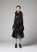 A woman with straight dark hair and a neutral expression stands against a plain gray background, wearing 132 5. ISSEY MIYAKE’s No.1-1: a sleeveless black recycled polyester dress with origami-like folds, shiny accents, and black ankle boots.