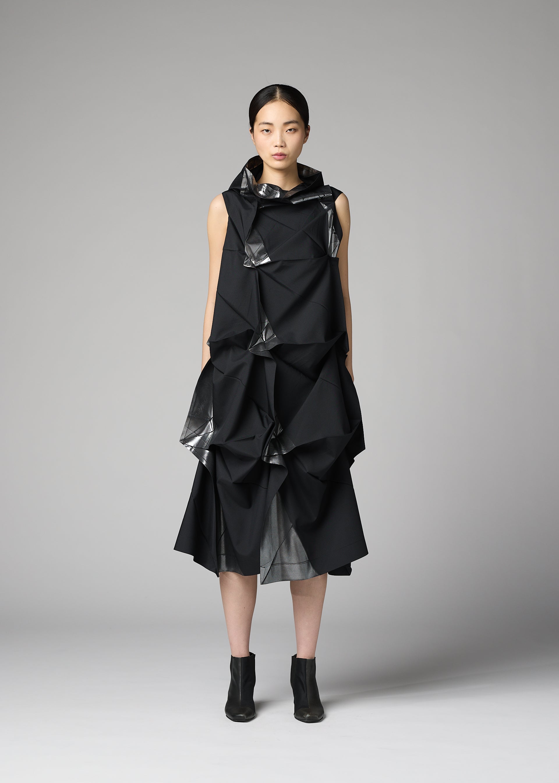Zoomed view A woman with straight dark hair and a neutral expression stands against a plain gray background, wearing 132 5. ISSEY MIYAKE’s No.1-1: a sleeveless black recycled polyester dress with origami-like folds, shiny accents, and black ankle boots.