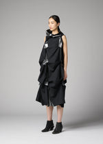 A woman models the 132 5. ISSEY MIYAKE No.1-1 dress—a sleeveless, knee-length design in recycled polyester with sculptural folds and metallic silver accents—paired with black ankle boots, set against a plain gray background.