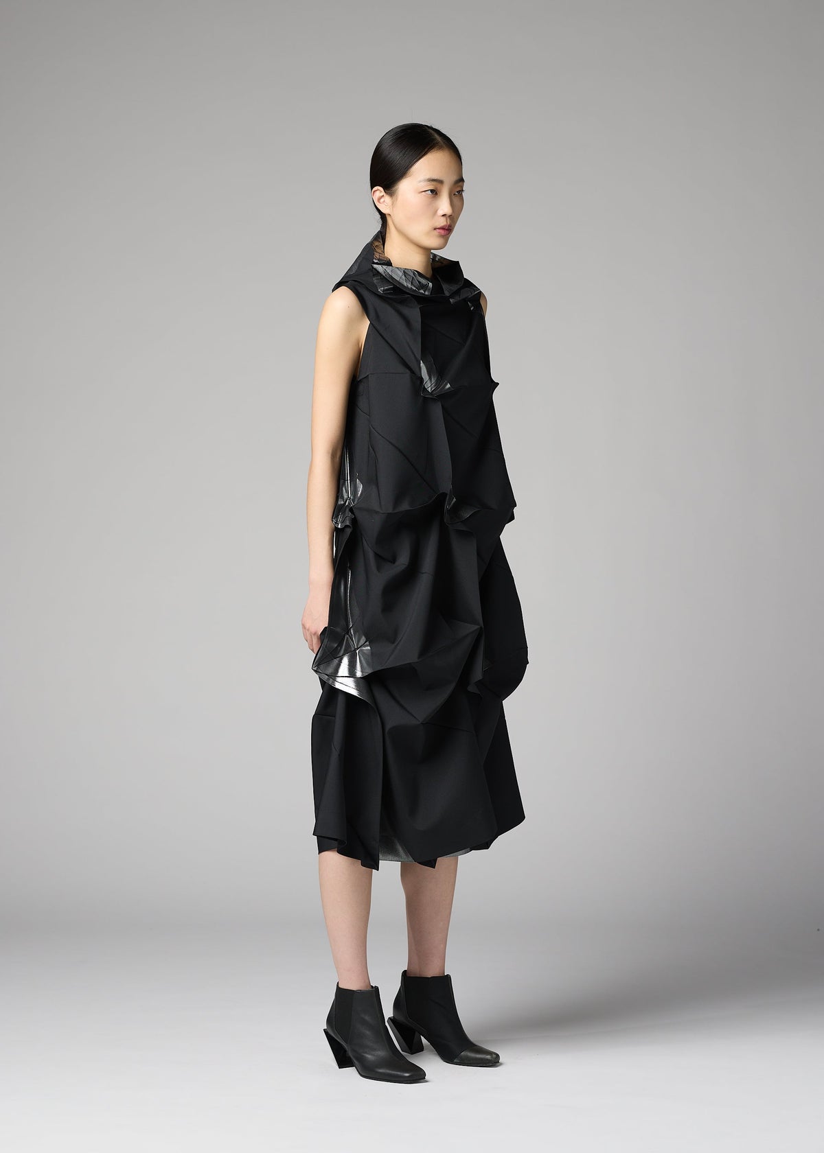 A woman with straight black hair stands in profile, wearing the 132 5. ISSEY MIYAKE No.1-1 sleeveless recycled polyester dress with sculptural layers and black ankle boots, set against a plain light gray background. view 8 of 9