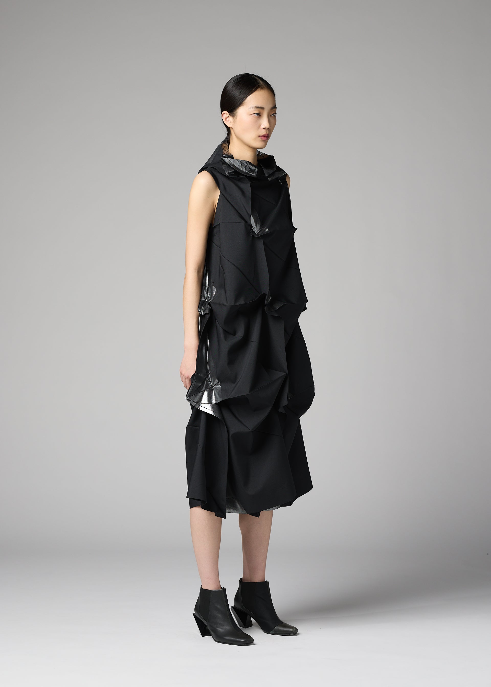 Zoomed view A woman with straight black hair stands in profile, wearing the 132 5. ISSEY MIYAKE No.1-1 sleeveless recycled polyester dress with sculptural layers and black ankle boots, set against a plain light gray background.