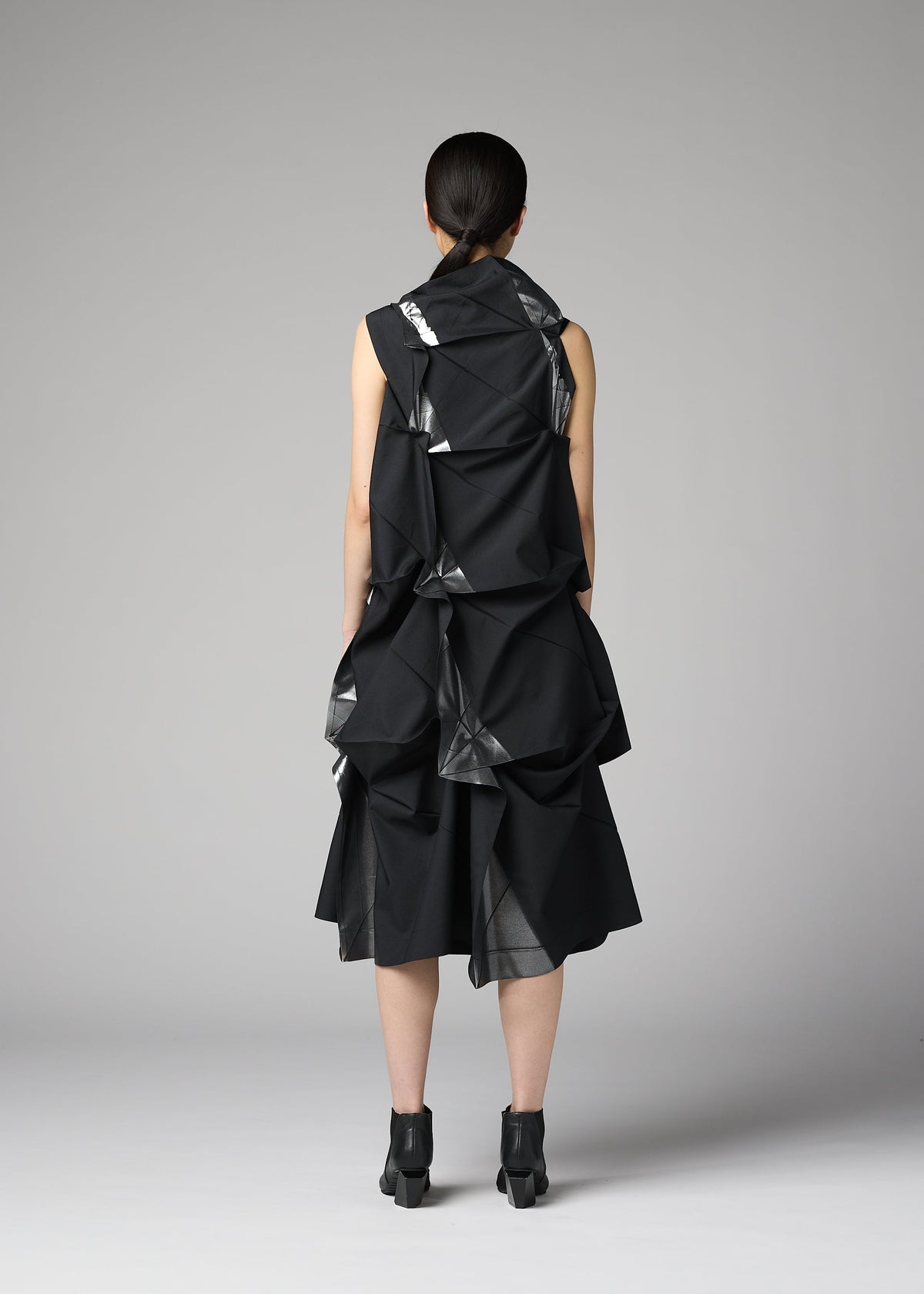 A woman with straight dark hair tied back wears the No.1-1 dress by 132 5. ISSEY MIYAKE, featuring recycled polyester, large geometric folds, and a pyramid silhouette, paired with black heels against a plain gray background. view 9 of 9