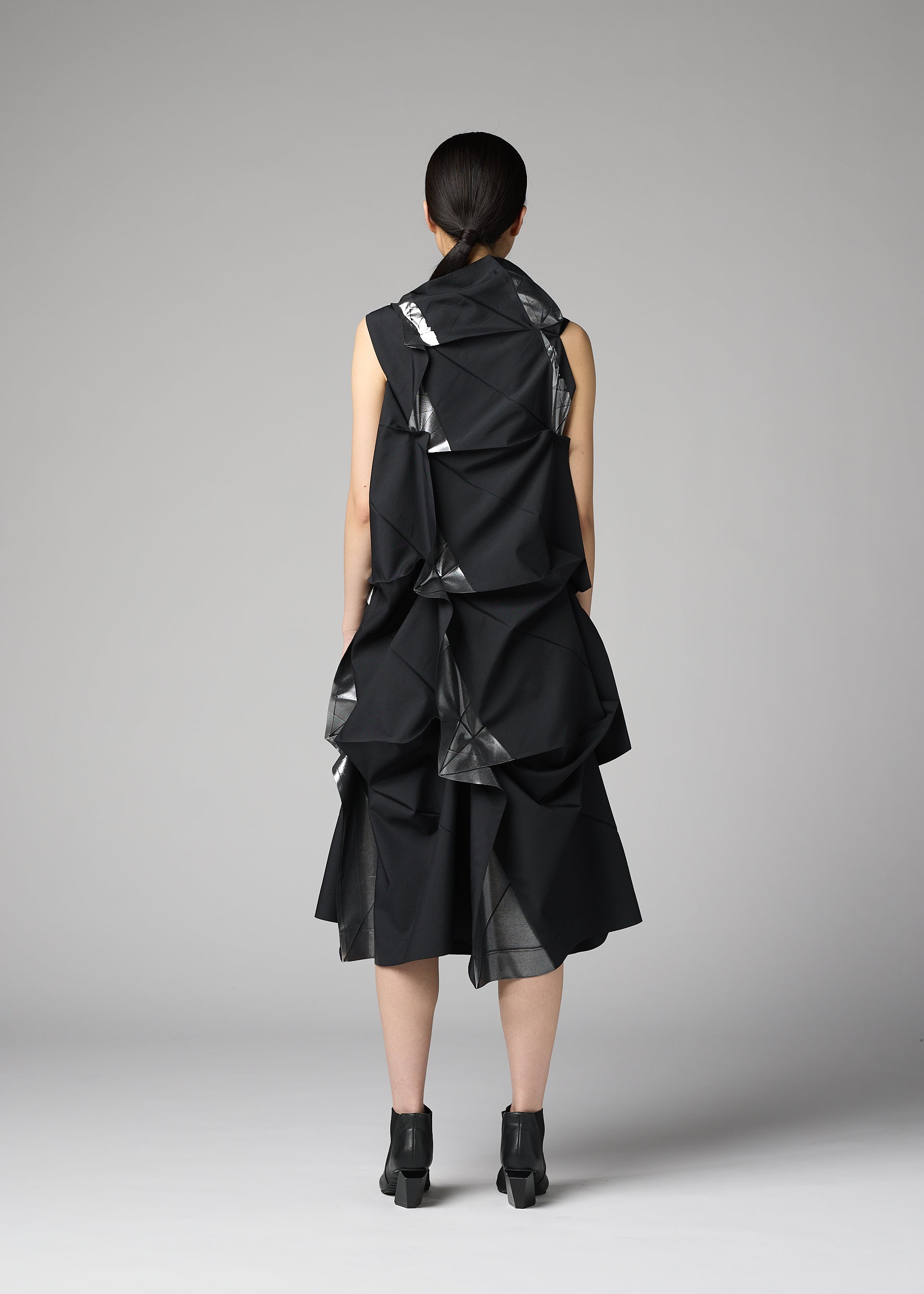 Zoomed view A woman with straight dark hair tied back wears the No.1-1 dress by 132 5. ISSEY MIYAKE, featuring recycled polyester, large geometric folds, and a pyramid silhouette, paired with black heels against a plain gray background.