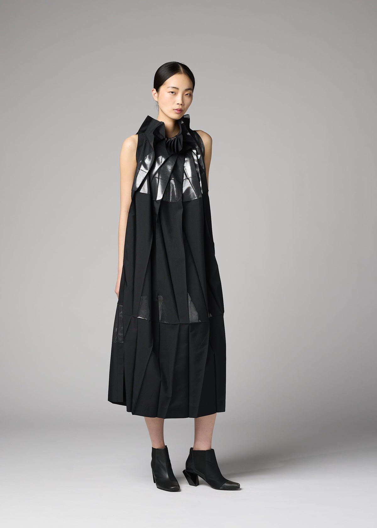 A woman stands against a gray background wearing the No.3-2 dress by 132 5. ISSEY MIYAKE—a sleeveless black recycled polyester piece with metallic accents, featuring overlapping folds and pleats—paired with black ankle boots. view 7 of 10