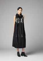 A woman stands against a gray background wearing the No.3-2 dress by 132 5. ISSEY MIYAKE—a sleeveless black recycled polyester piece with metallic accents, featuring overlapping folds and pleats—paired with black ankle boots.