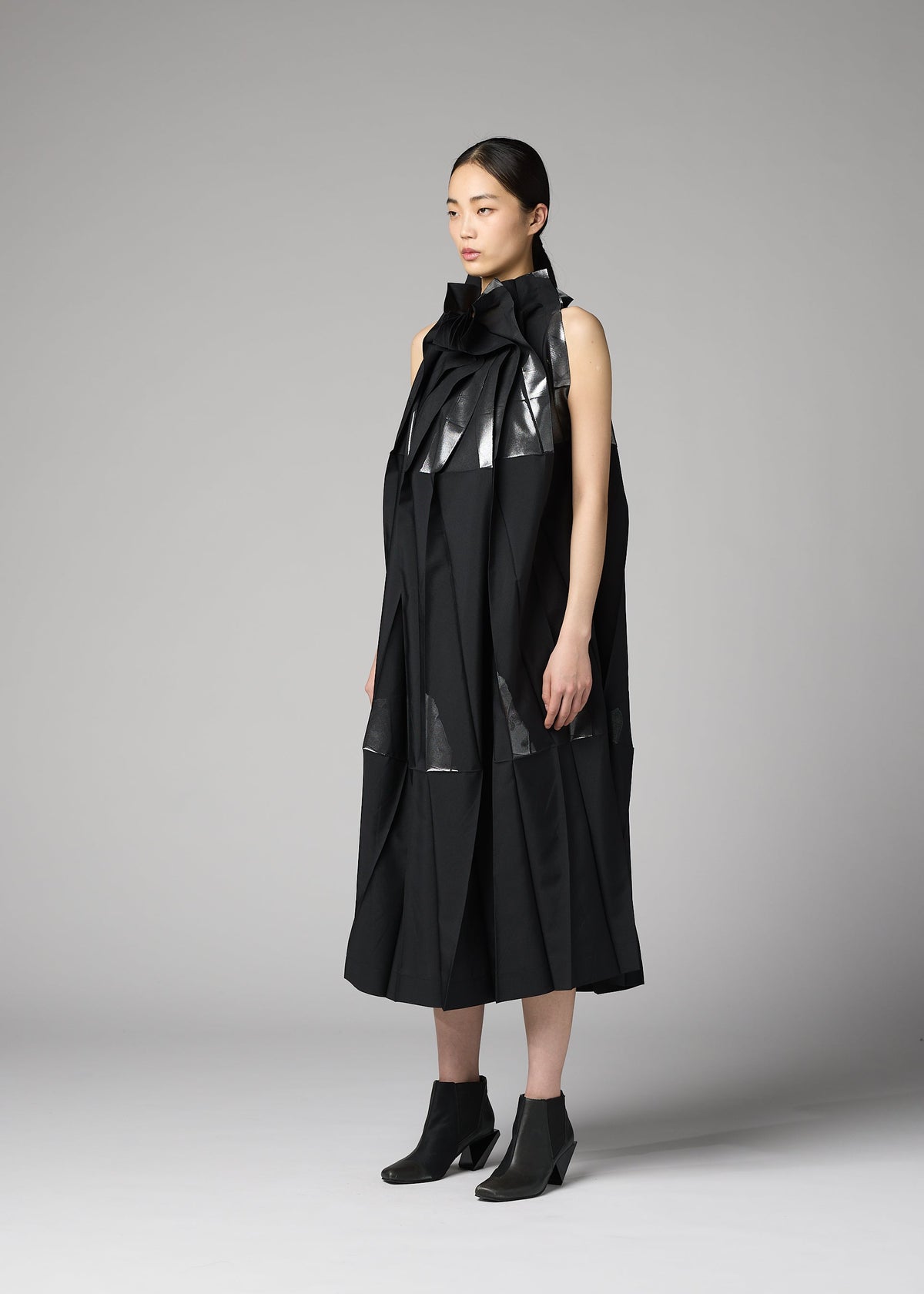 A woman stands in a studio wearing the 132 5. ISSEY MIYAKE No.3-2 dress, a black, sleeveless piece crafted from recycled polyester with sculptural, glossy accents and overlapping folds inspired by foil processing techniques. Paired with black ankle boots, the modern, architectural design lends her look a distinctive edge as she faces slightly to the side with a neutral expression. view 8 of 10