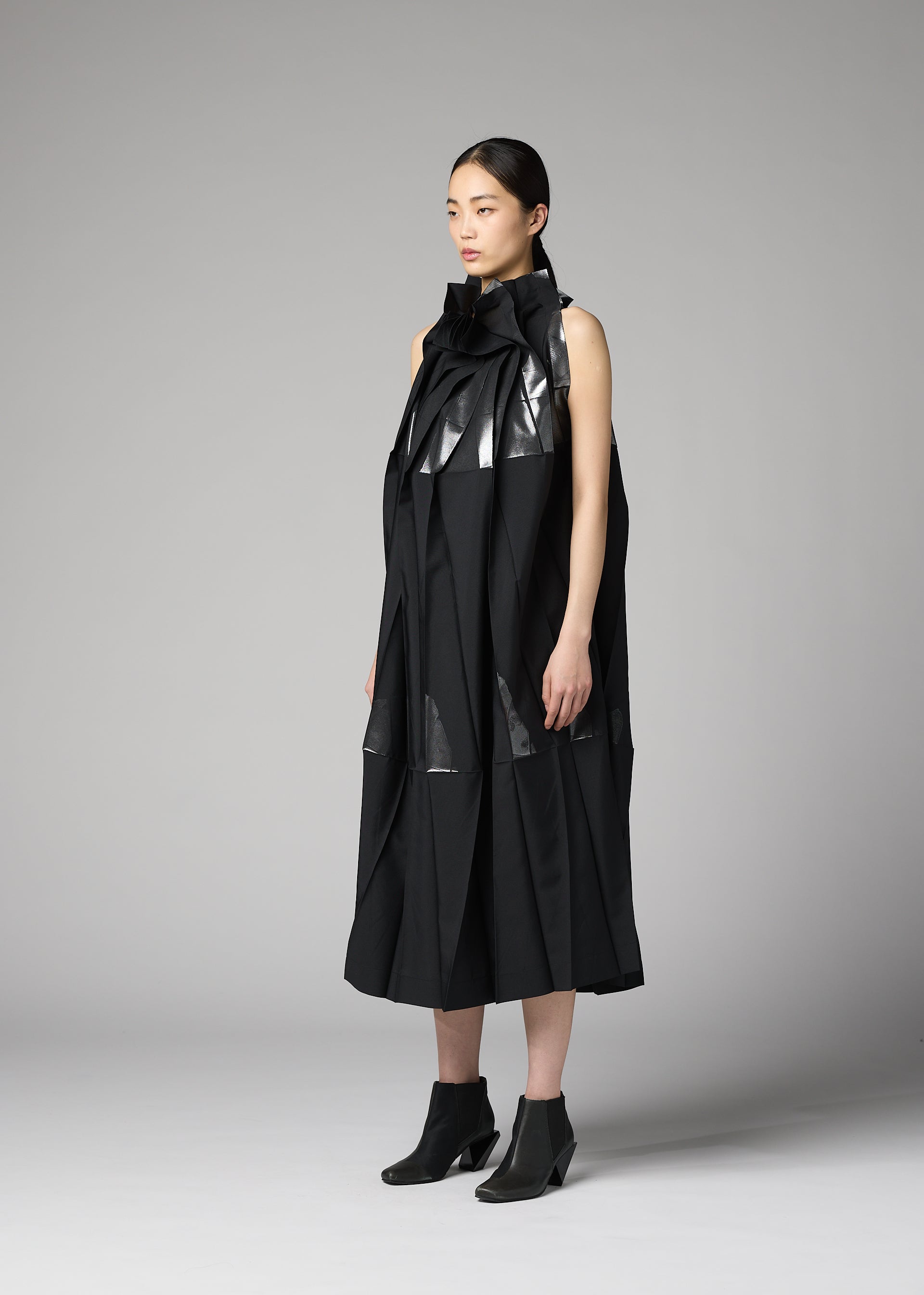 Zoomed view A woman stands in a studio wearing the 132 5. ISSEY MIYAKE No.3-2 dress, a black, sleeveless piece crafted from recycled polyester with sculptural, glossy accents and overlapping folds inspired by foil processing techniques. Paired with black ankle boots, the modern, architectural design lends her look a distinctive edge as she faces slightly to the side with a neutral expression.