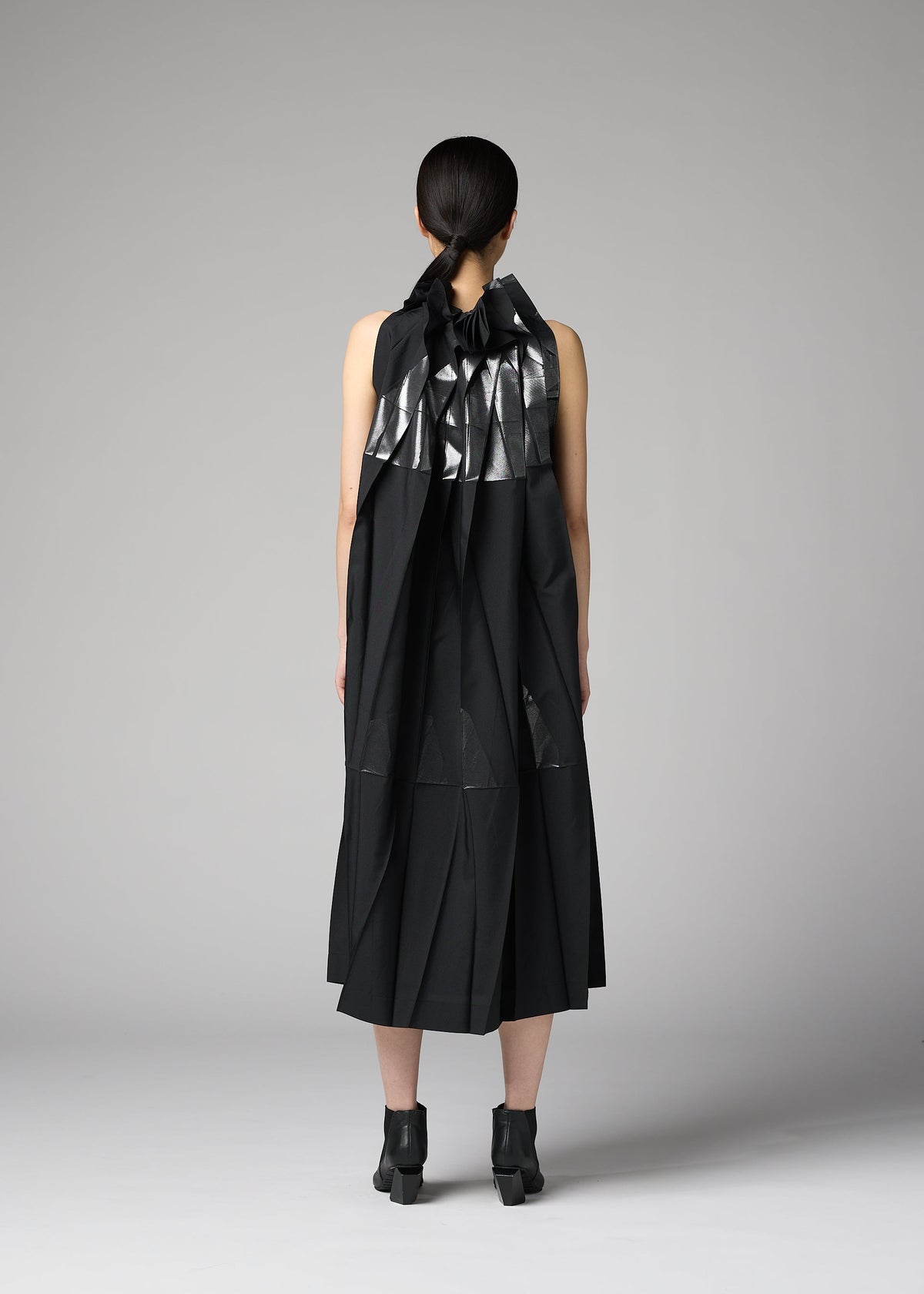 A person with dark hair tied back faces away, wearing the No.3-2 dress by 132 5. ISSEY MIYAKE—a sleeveless black recycled polyester dress featuring metallic silver geometric accents and overlapping folds—paired with black ankle boots, standing against a plain gray background. view 9 of 10