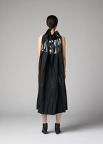 A person with dark hair tied back faces away, wearing the No.3-2 dress by 132 5. ISSEY MIYAKE—a sleeveless black recycled polyester dress featuring metallic silver geometric accents and overlapping folds—paired with black ankle boots, standing against a plain gray background.