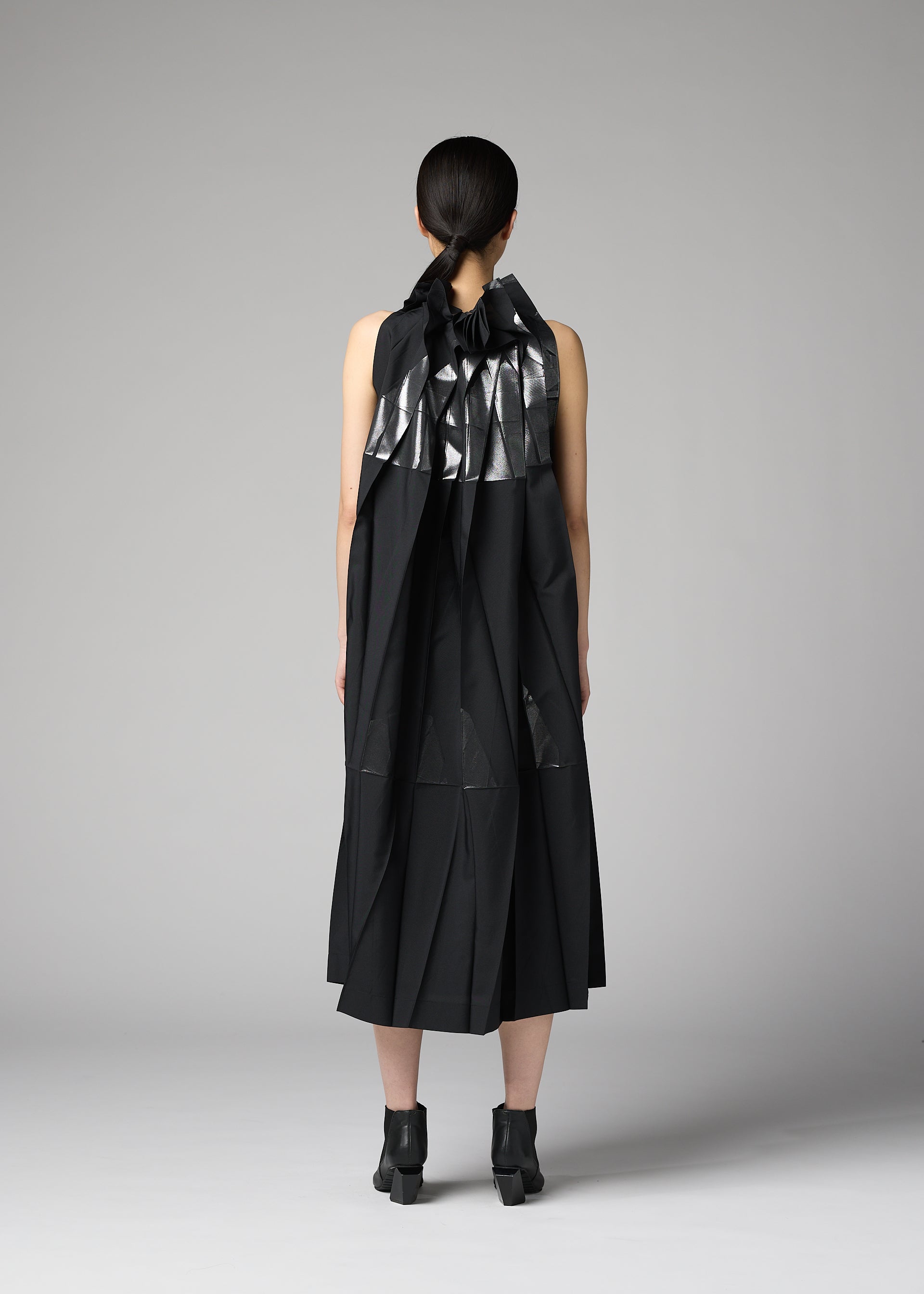Zoomed view A person with dark hair tied back faces away, wearing the No.3-2 dress by 132 5. ISSEY MIYAKE—a sleeveless black recycled polyester dress featuring metallic silver geometric accents and overlapping folds—paired with black ankle boots, standing against a plain gray background.
