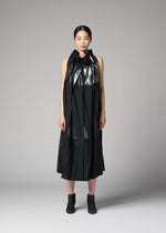 A person stands facing forward against a plain gray background, wearing the No.3-2 dress by 132 5. ISSEY MIYAKE— a black, sleeveless recycled polyester dress featuring overlapping folds and a shiny ruffled collar—paired with black ankle boots.