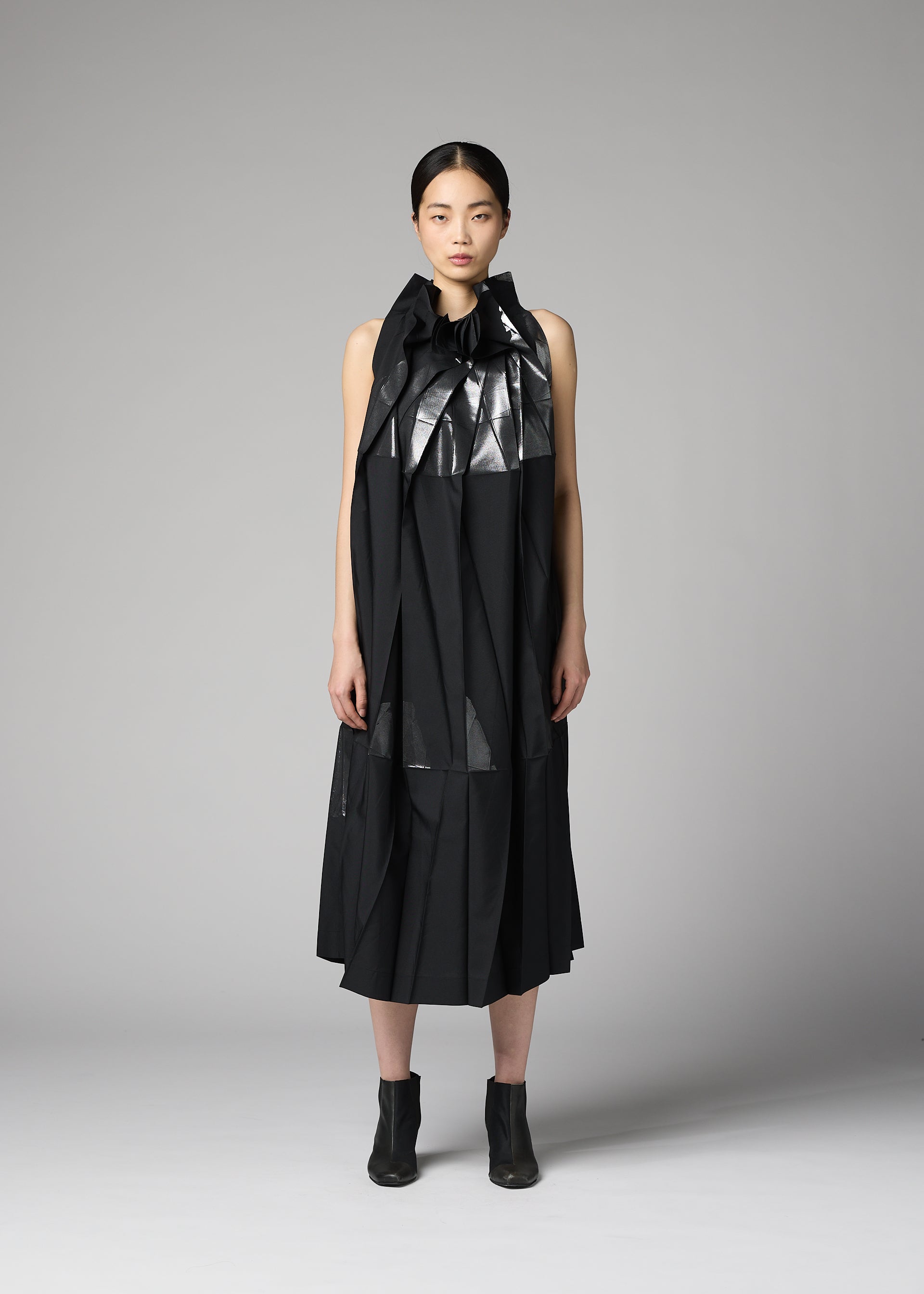 Zoomed view A person stands facing forward against a plain gray background, wearing the No.3-2 dress by 132 5. ISSEY MIYAKE— a black, sleeveless recycled polyester dress featuring overlapping folds and a shiny ruffled collar—paired with black ankle boots.