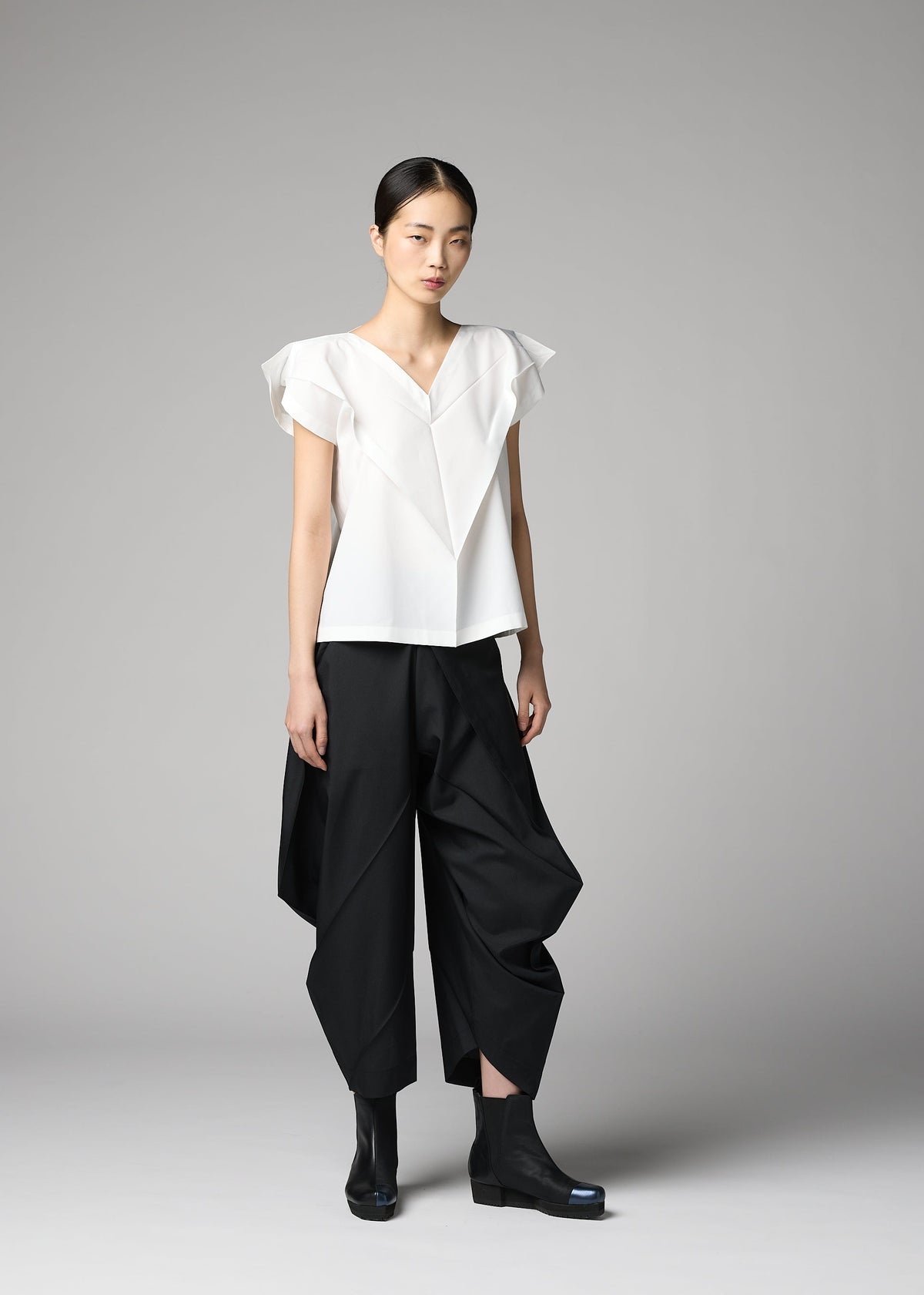 A woman with straight dark hair pulled back stands against a gray background wearing the 132 5. ISSEY MIYAKE F-SH-1 white folding technique shirt with ruffle details, loose black origami-style pants, and black ankle boots. view 6 of 8