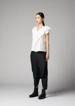 A woman stands against a plain gray background wearing the F-SH-1 shirt by 132 5. ISSEY MIYAKE, loose black draped pants, and black ankle boots. She faces forward with a neutral expression.
