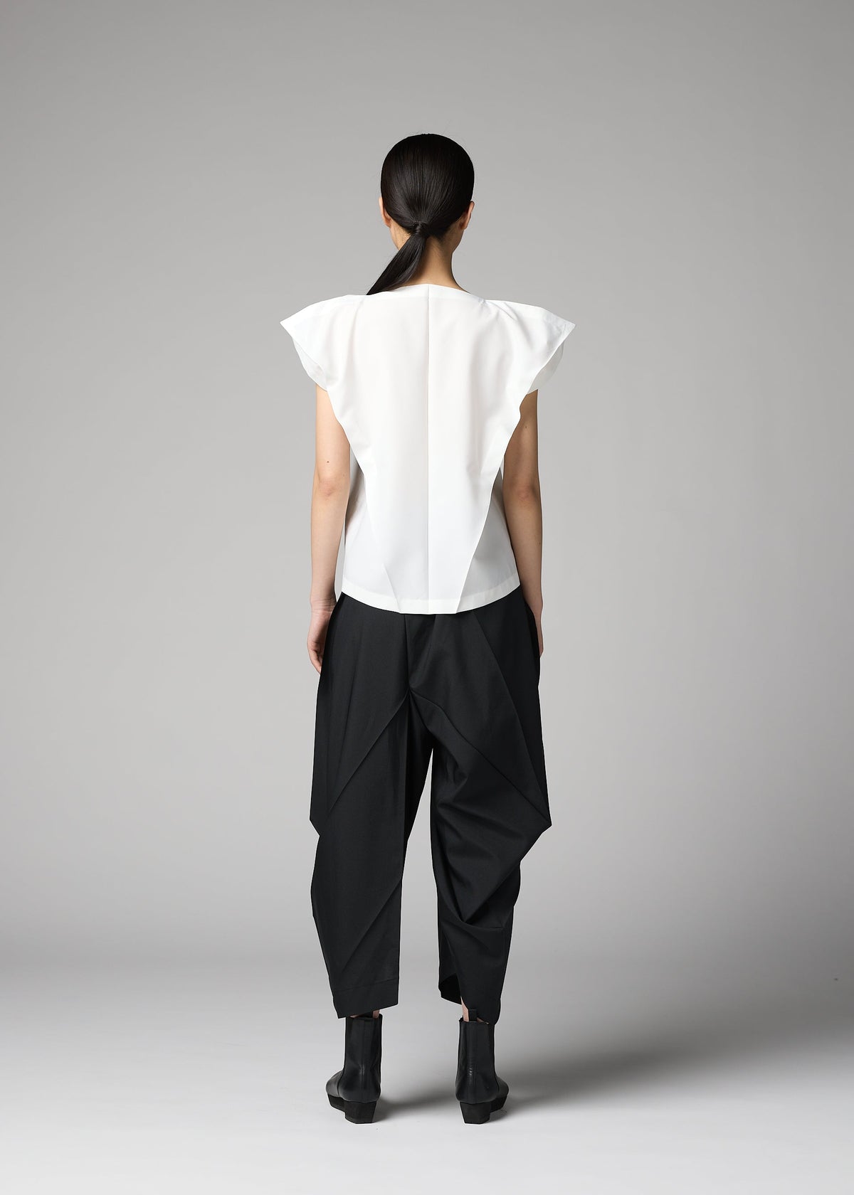 A person with long dark hair tied back stands facing away, wearing the F-SH-1 folding technique shirt by 132 5. ISSEY MIYAKE, pointed shoulders visible, paired with loose black geometric pants and black ankle boots against a gray background. view 8 of 8