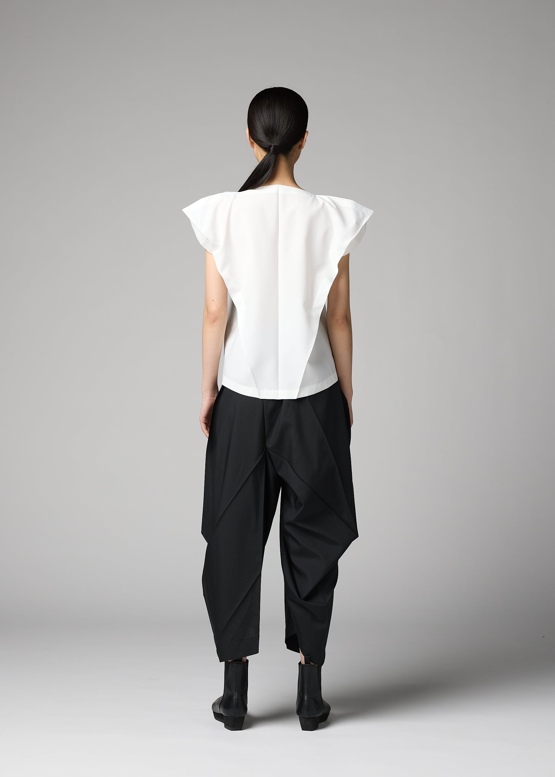 Zoomed view A person with long dark hair tied back stands facing away, wearing the F-SH-1 folding technique shirt by 132 5. ISSEY MIYAKE, pointed shoulders visible, paired with loose black geometric pants and black ankle boots against a gray background.