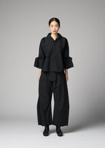 A woman stands in a studio wearing 132 5. ISSEY MIYAKE’s F-SH-1: a structured, black, geometric-patterned outfit with wide trousers and a loose recycled polyester shirt. Her hair is pulled back, and she faces forward neutrally.