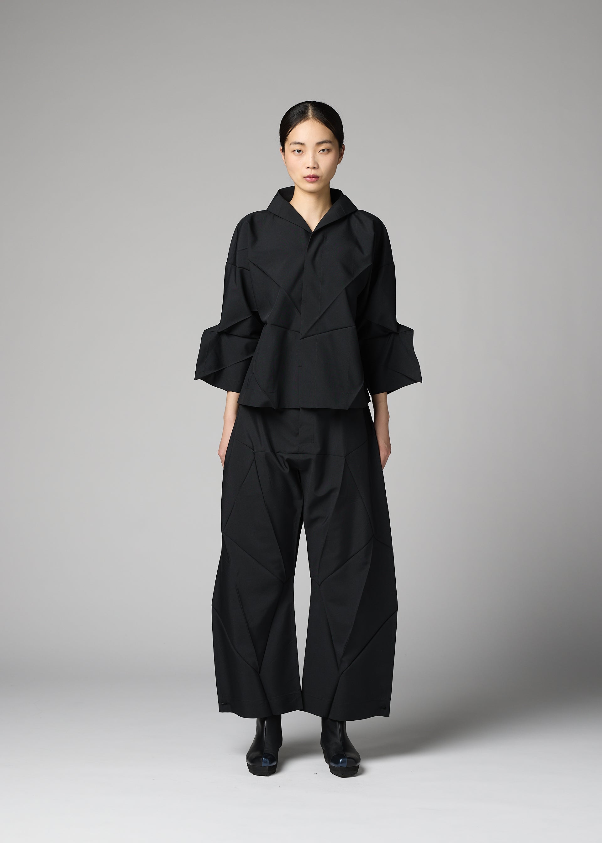 Zoomed view A woman stands in a studio wearing 132 5. ISSEY MIYAKE’s F-SH-1: a structured, black, geometric-patterned outfit with wide trousers and a loose recycled polyester shirt. Her hair is pulled back, and she faces forward neutrally.