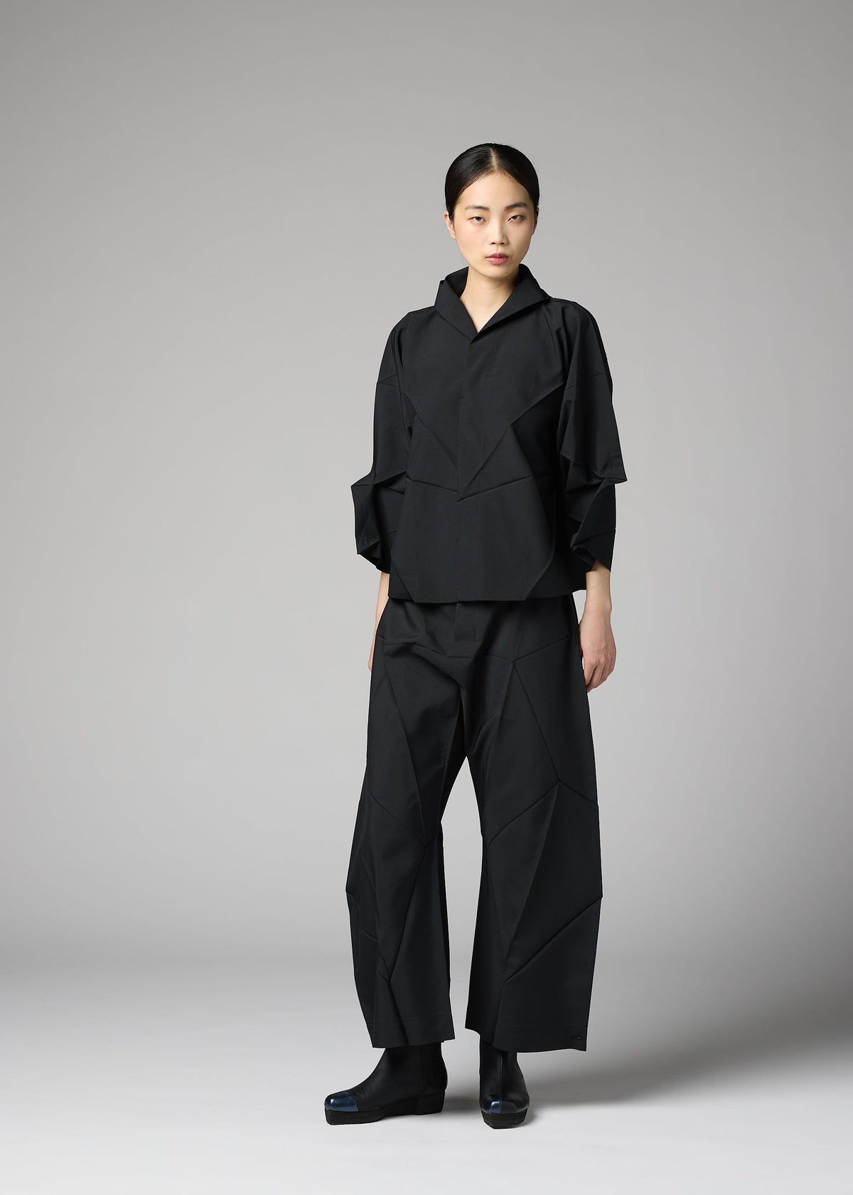 A woman with her hair tied back and a neutral expression stands before a gray backdrop, wearing the F-SH-1 folding technique shirt by 132 5. ISSEY MIYAKE, paired with wide-leg pants and matching black shoes. view 7 of 8