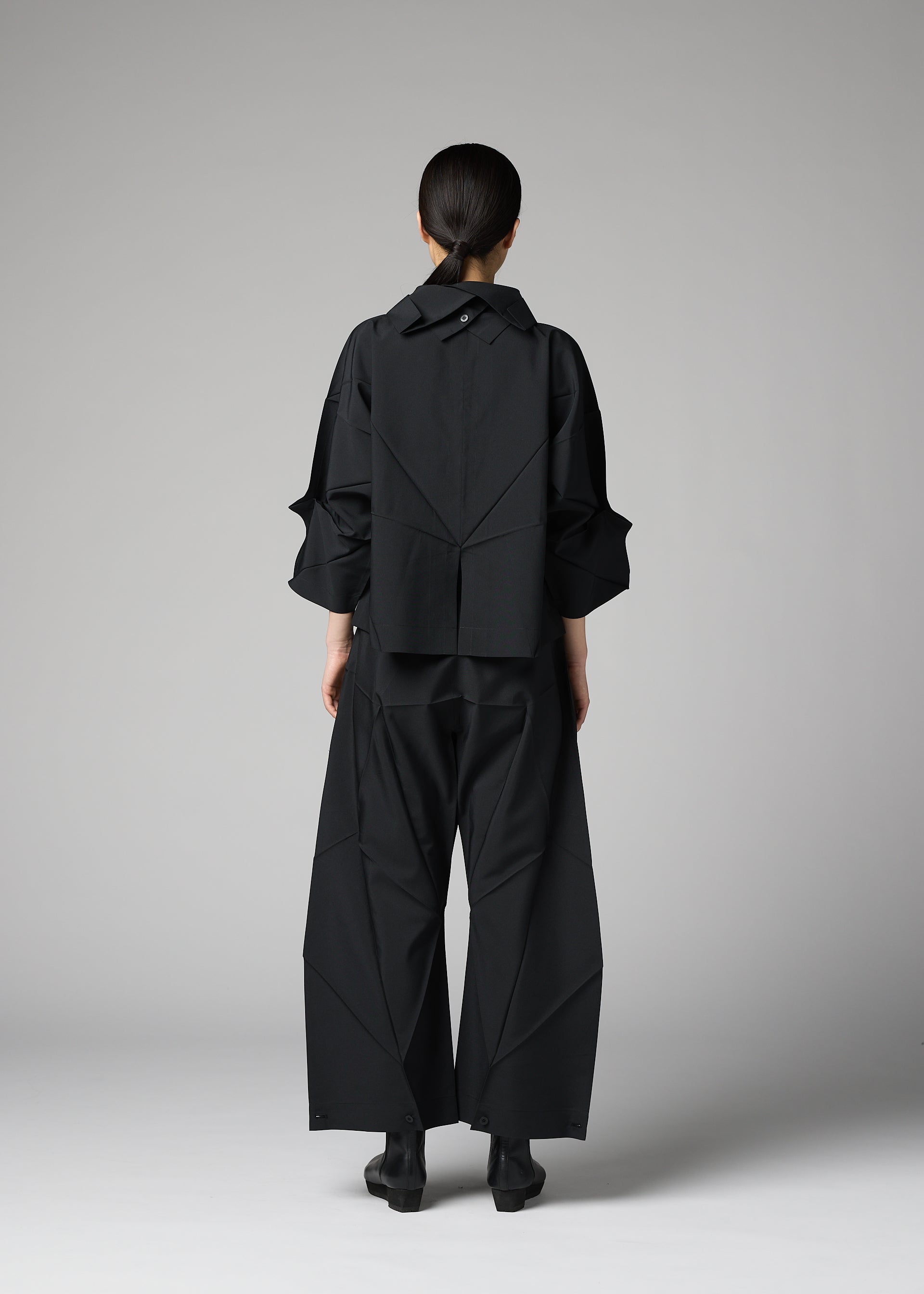 Zoomed view A person stands facing away, wearing the F-SH-1 shirt from 132 5. ISSEY MIYAKE. The light gray background highlights the unique geometric folds and structured design of this loose-fitting black piece.