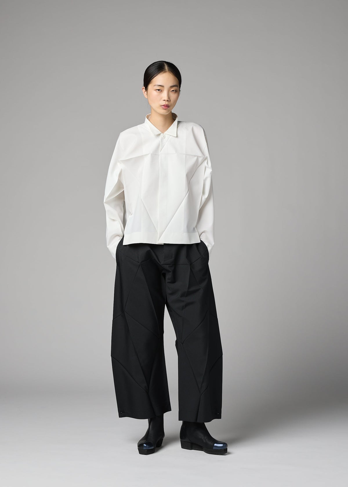 A person stands against a gray background wearing the 132 5. ISSEY MIYAKE F-SH-2 shirt, paired with wide black pants and black shoes. They have a neutral expression and straight, dark hair pulled back. view 9 of 12