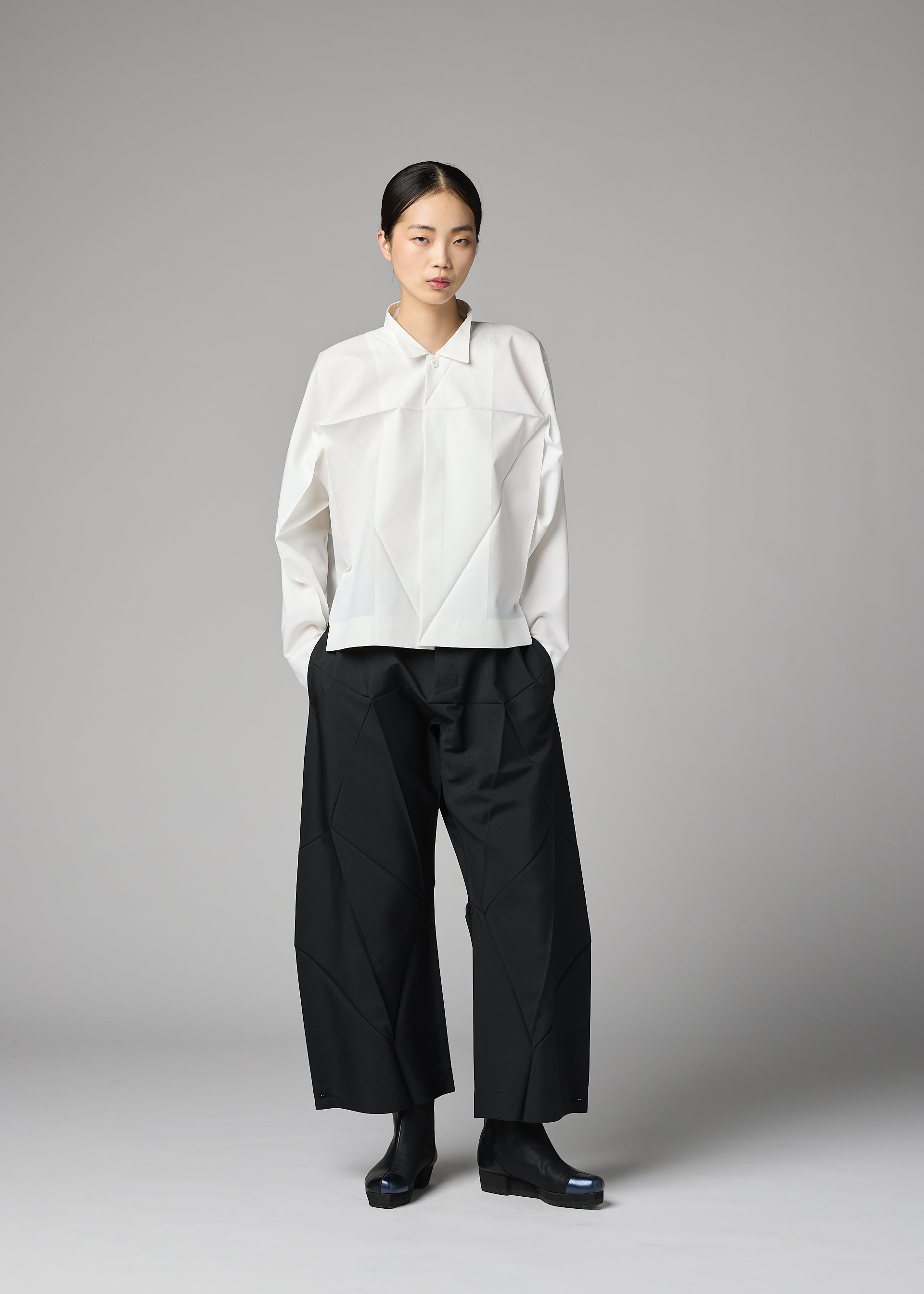 Zoomed view A person stands against a gray background wearing the 132 5. ISSEY MIYAKE F-SH-2 shirt, paired with wide black pants and black shoes. They have a neutral expression and straight, dark hair pulled back.