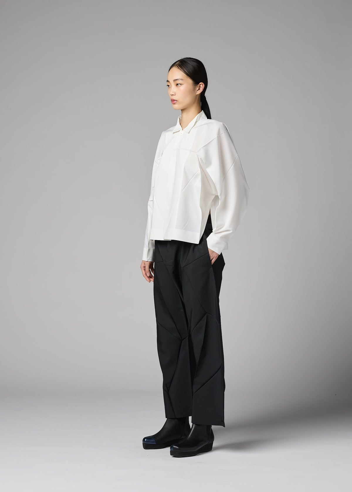 A person stands in a studio against a plain gray background, wearing the F-SH-2 loose white recycled polyester shirt with a standing collar from 132 5. ISSEY MIYAKE, paired with black wide-leg pants and black boots, and maintains a neutral expression. view 10 of 12