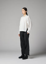 A person stands in a studio against a plain gray background, wearing the F-SH-2 loose white recycled polyester shirt with a standing collar from 132 5. ISSEY MIYAKE, paired with black wide-leg pants and black boots, and maintains a neutral expression.
