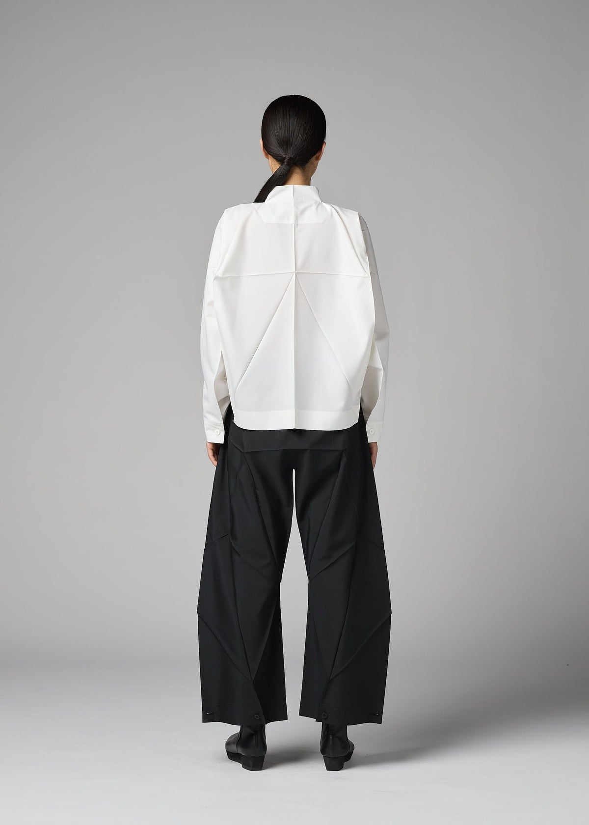 A person stands facing away, wearing the 132 5. ISSEY MIYAKE F-SH-2 shirt featuring geometric folds and wide-leg black pants. Their hair is tied back, set against a plain gray background. view 11 of 12